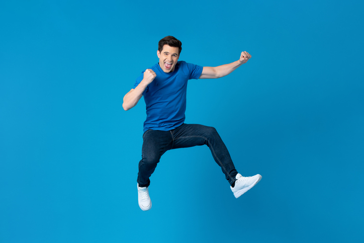Smiling handsome American man joyfully jumping and raising his fists isolated on blue studio background fro success and freedom concepts