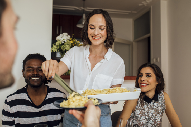 Pretty smiling Caucasian woman hosting dinner party at home and putting pasta on friends plate.